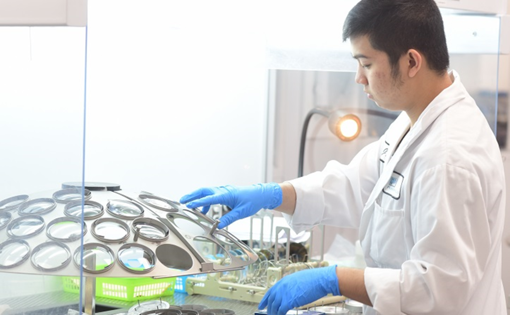 Optical lab technician in blue gloves inspecting a tray of lenses inside a clean bench during coating/quality control.