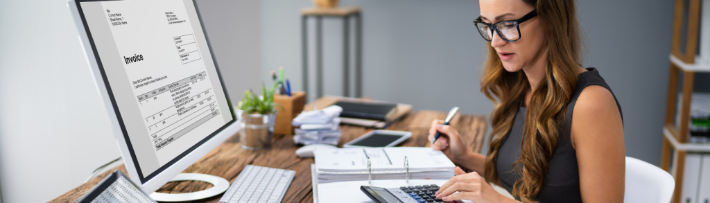 An image of a woman with glasses using a calculator and with a laptop in front of her.