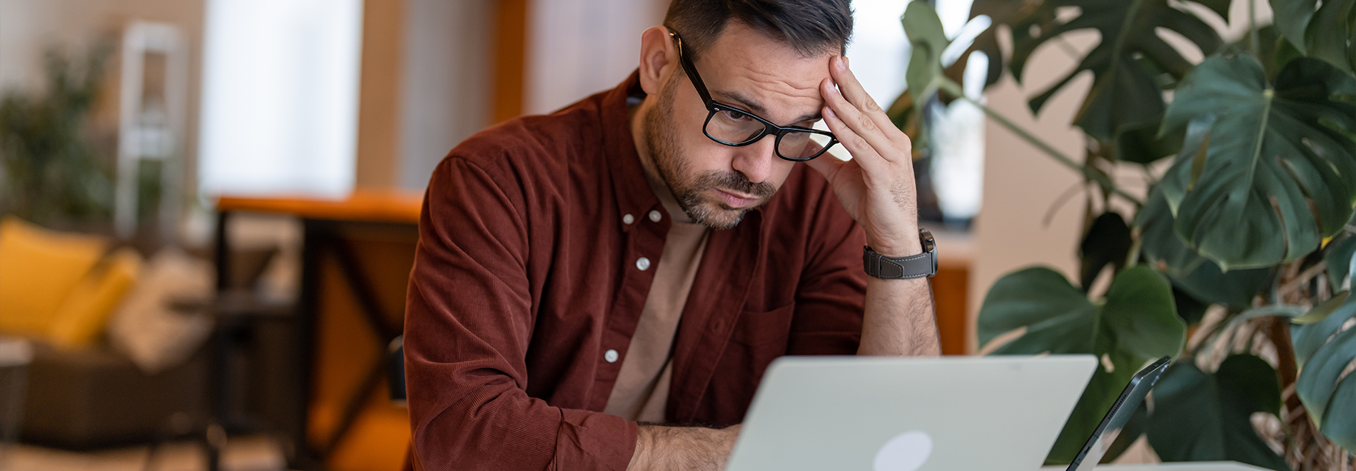 Stressed man wearing glasses sitting at a desk with a hand on his forehead, looking at a laptop screen in a home office with plants in the background."