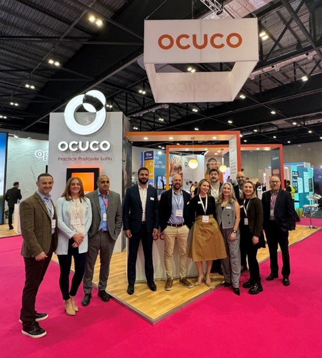 Alt text: A group of Ocuco employees standing together in front of the Ocuco booth at 100% Optical 2024 2024. The booth has the Ocuco logo displayed prominently on a tall structure with orange accents. Everyone is smiling and dressed in business attire, standing on a pink carpeted exhibition floor.