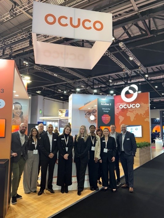 A team of Ocuco employees posing in front of the Ocuco exhibition booth at 100% Optical 2025. The booth features branding for Acuitas 3 and Ocuco’s global presence, with orange and gray design elements. The group is smiling and wearing business attire on a wooden platform with black carpeted walkways.