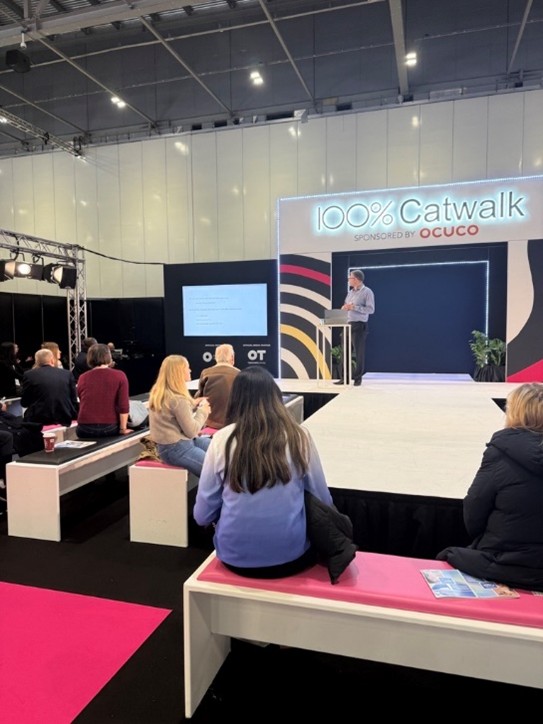 An Ocuco-sponsored Catwalk stage during 100% Optical 2025. Attendees are seated on benches watching a speaker present slides on a large screen. The stage backdrop reads “100% Catwalk Sponsored by Ocuco” in illuminated text, with bold black, white, and pink design elements.