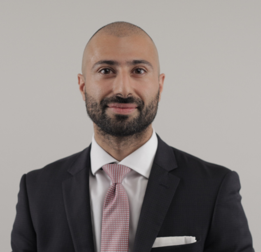 Kishan Devraj, UK-qualified optometrist, wearing a dark suit with a white shirt and light red patterned tie, smiling against a plain light background.