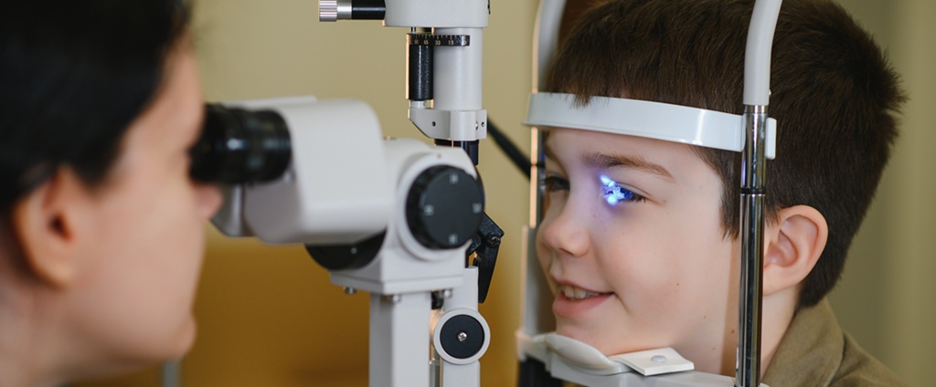 A young boy smiles while having his eyes examined with a slit lamp by an eye care professional during a clinical assessment.